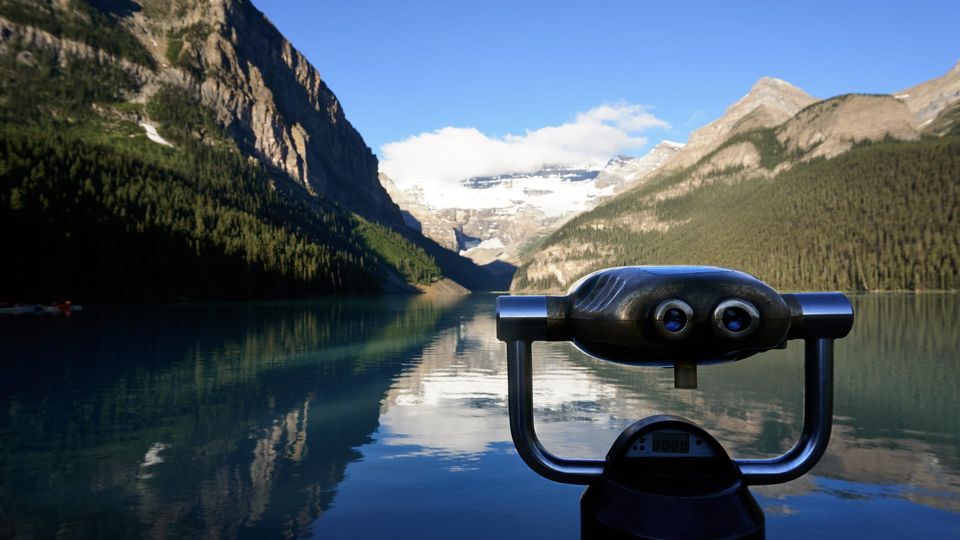 Coin-operated binoculars in front of Lake Louise reflecting a glacier and forested mountains