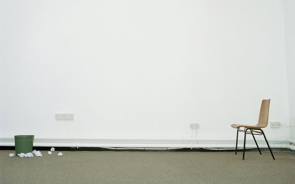 Empty white room with a lone wooden chair and a green wastebasket surrounded by crumpled paper