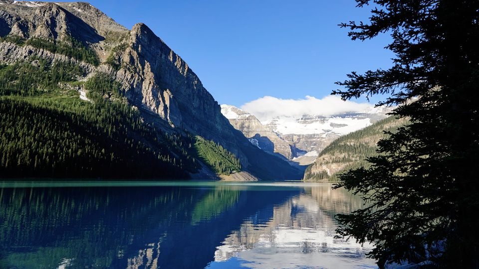 Lake Louise with steep forested mountains and a snowy glacier reflected in calm blue water, framed by a dark evergreen