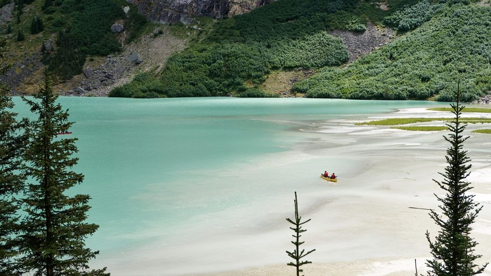 Two people in red vests paddling a yellow canoe across a turquoise glacial lake below green mountain slopes