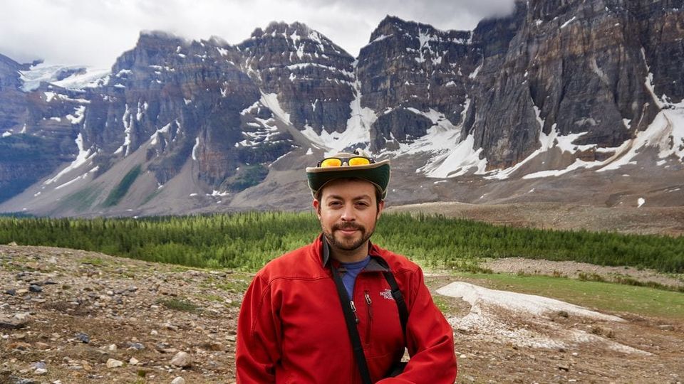 Sam in a red jacket and wide-brim hat with sunglasses in front of snowy rugged mountains