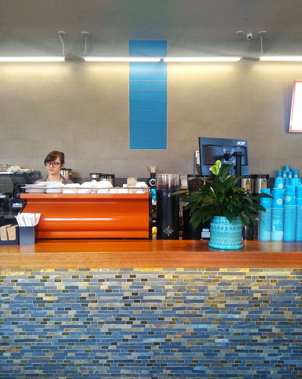 Barista in glasses behind an orange espresso machine at a tiled counter with stacks of blue cups
