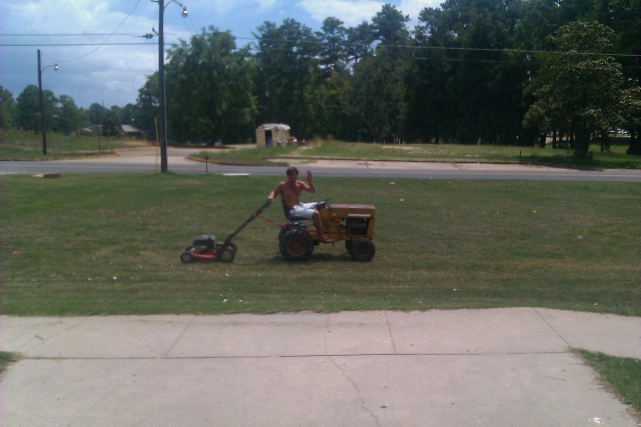 A shirtless man rides a small yellow tractor while towing a push mower across a grassy lawn, waving at the camera