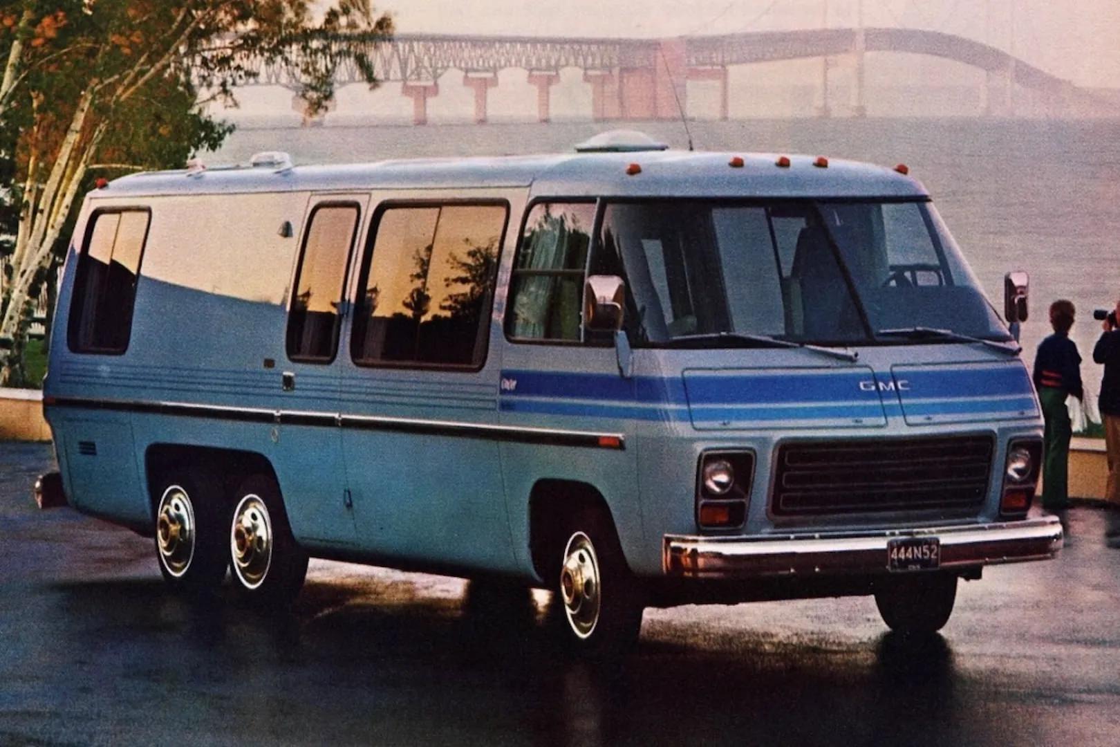 Vintage blue GMC motorhome with a six-wheel configuration parked near a waterway with a large bridge visible in the misty background
