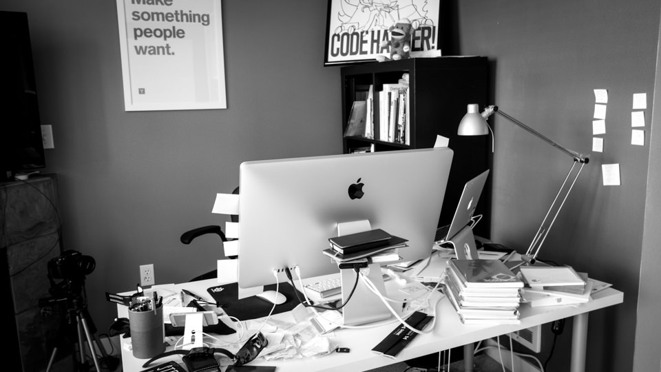 A cluttered home office desk with an iMac, laptop, books, and a lamp, with "Make something people want" and "Code Harder" posters on the wall