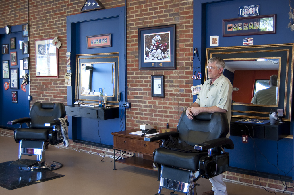 A man stands beside a barber chair in a barbershop with brick walls, blue trim, mirrors and sports memorabilia including a "War Eagle" sign