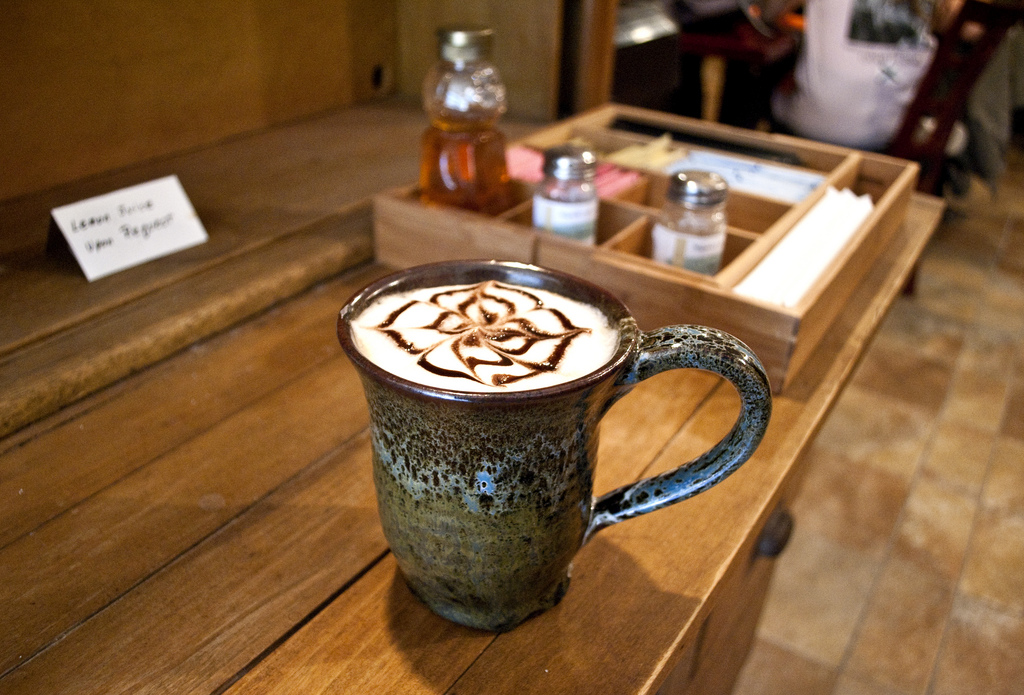 A latte with chocolate syrup art in a handmade ceramic mug on a wooden table, with a condiment caddy and honey jar in the background