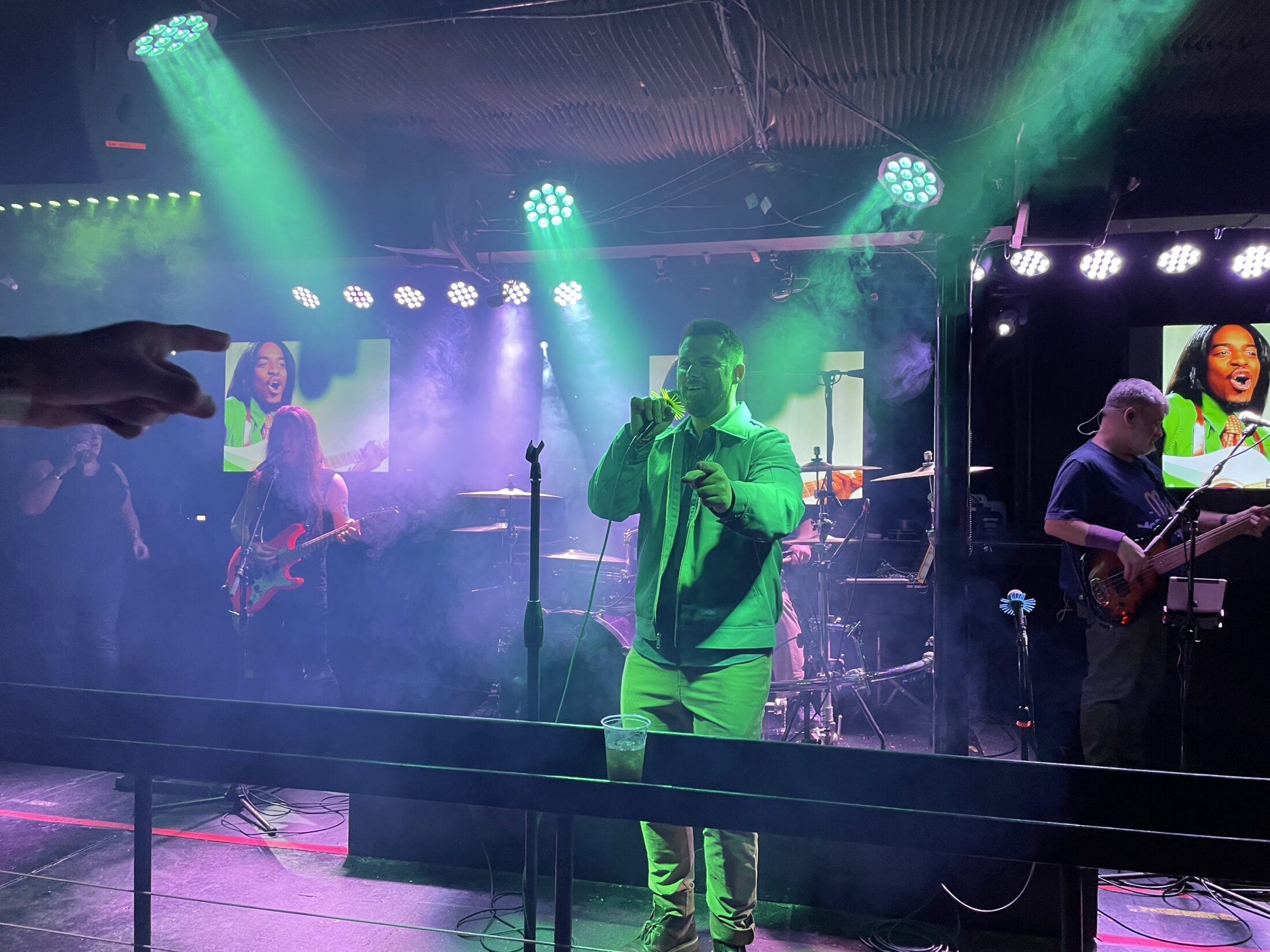 A live band performs on a smoke-filled stage under green lights, with a male vocalist holding a microphone, a guitarist, and a bassist visible