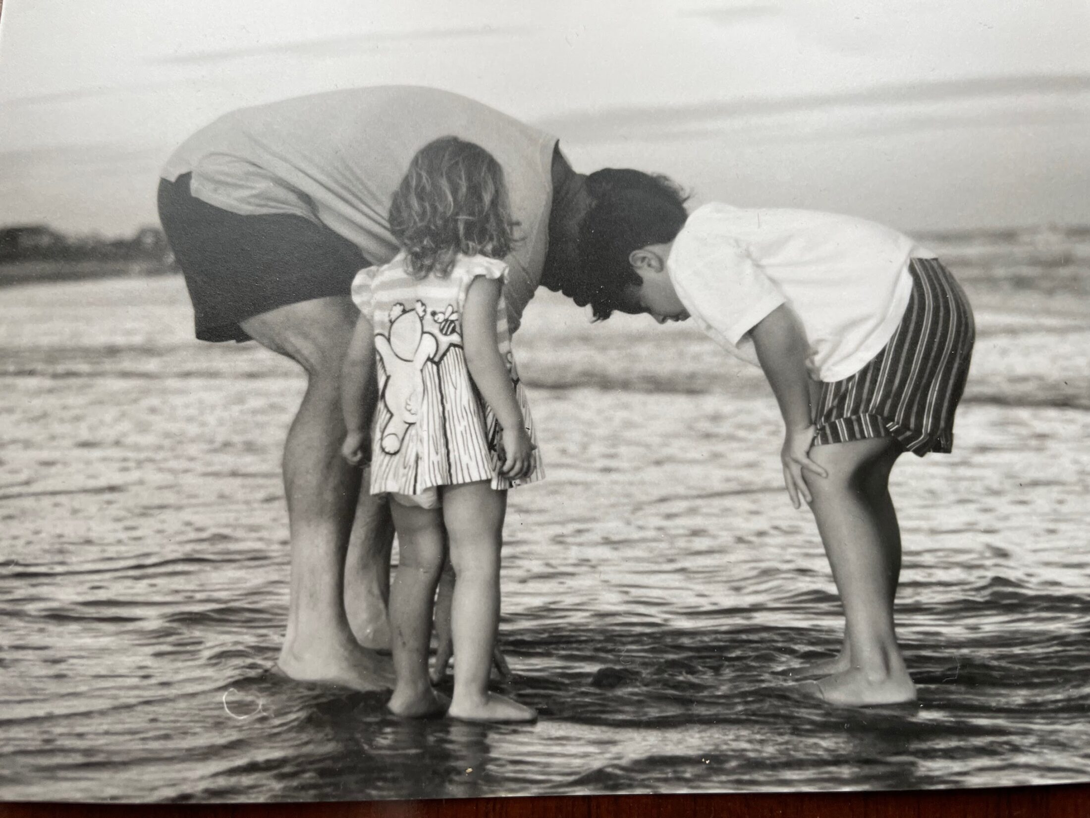 Vintage black-and-white photo of an adult and two young children bending down to look at the water's edge on a beach