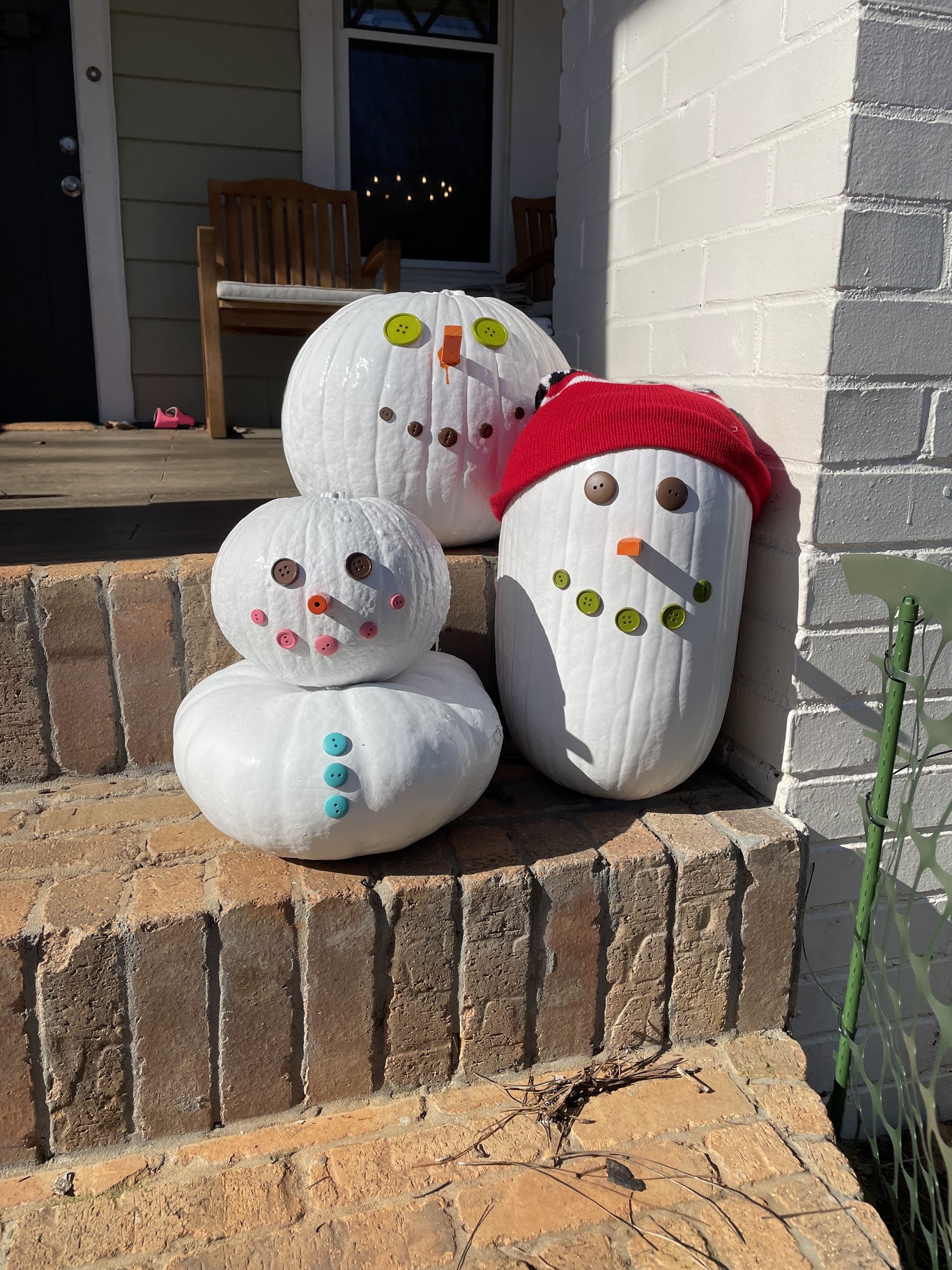 Three white-painted pumpkins decorated as snowmen with button eyes, orange noses, and colorful button accents, sitting on brick porch steps