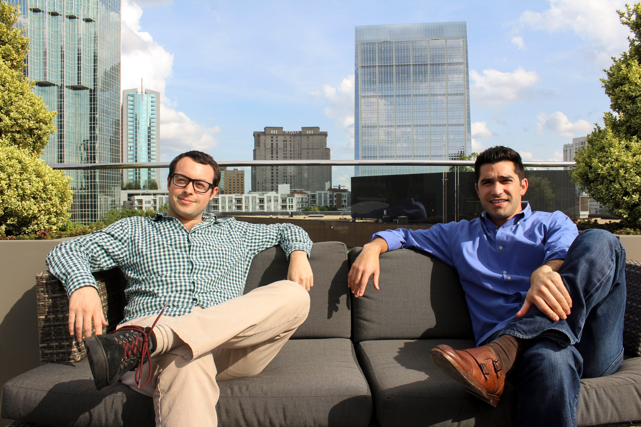 Two men sitting on a rooftop couch with a city skyline behind them on a sunny day