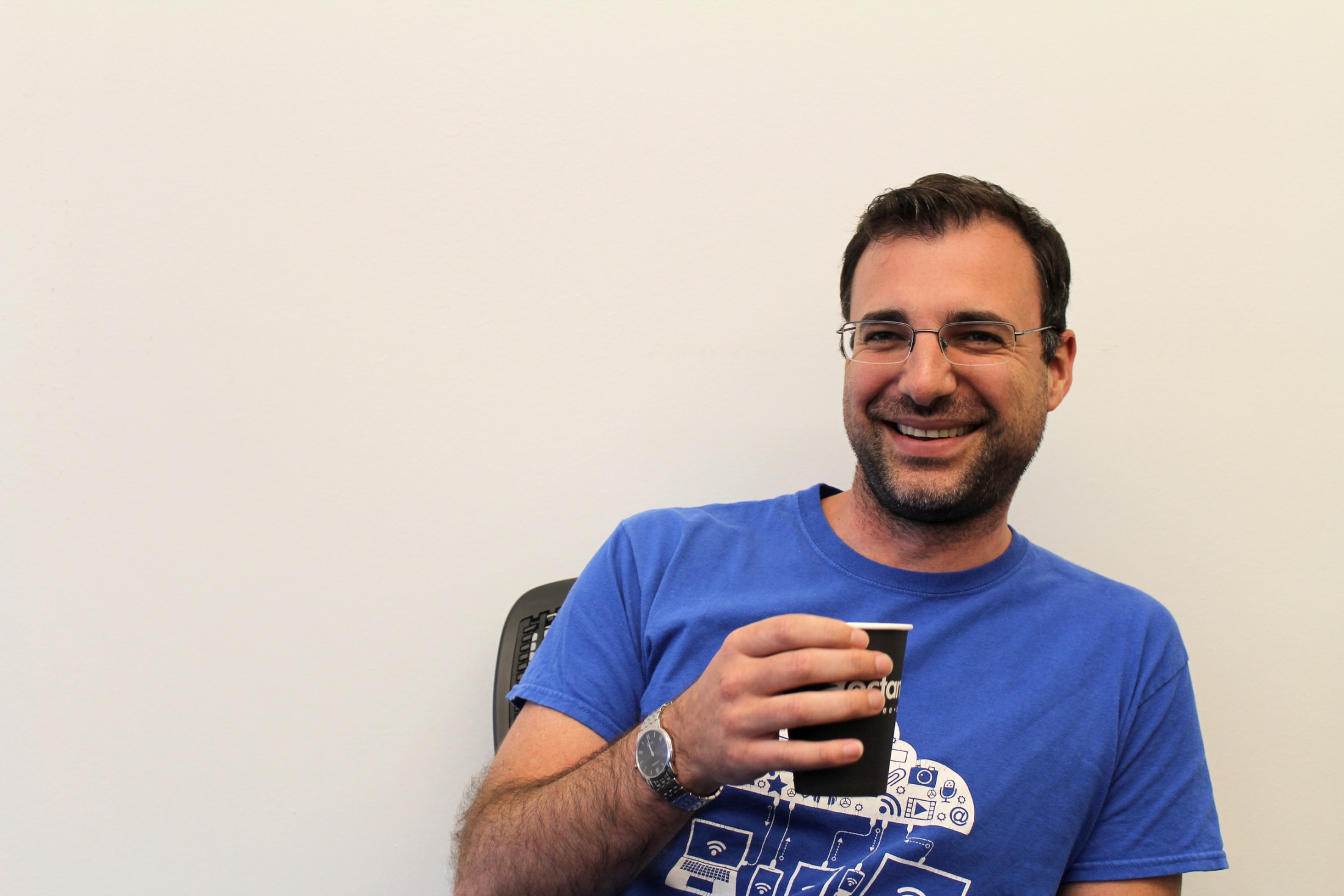 A smiling man with dark hair, a short beard, and thin-framed glasses holding a black paper cup while seated in an office chair
