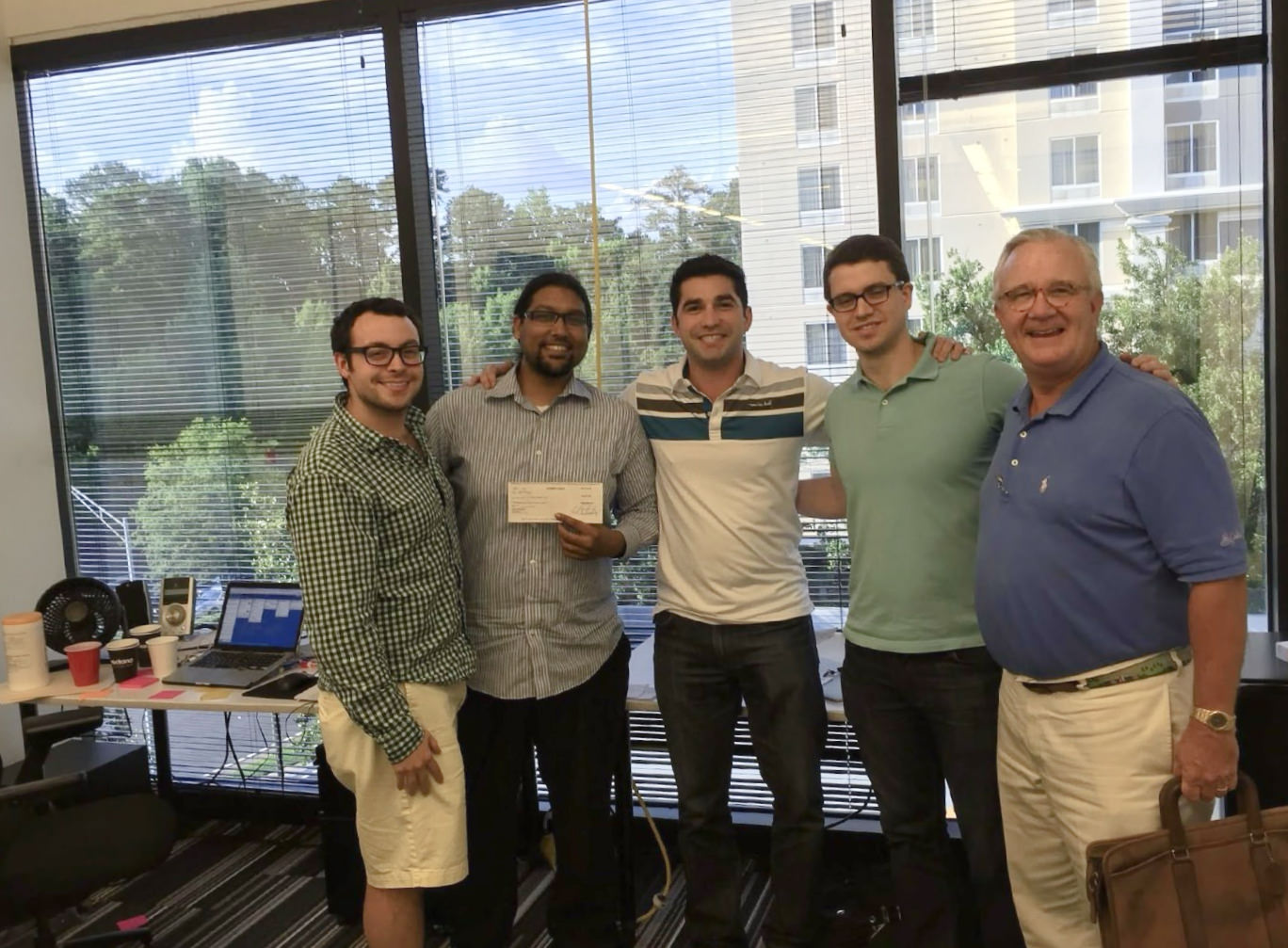 Five men posing together indoors, one holding a check, with a laptop and office supplies on a table behind them
