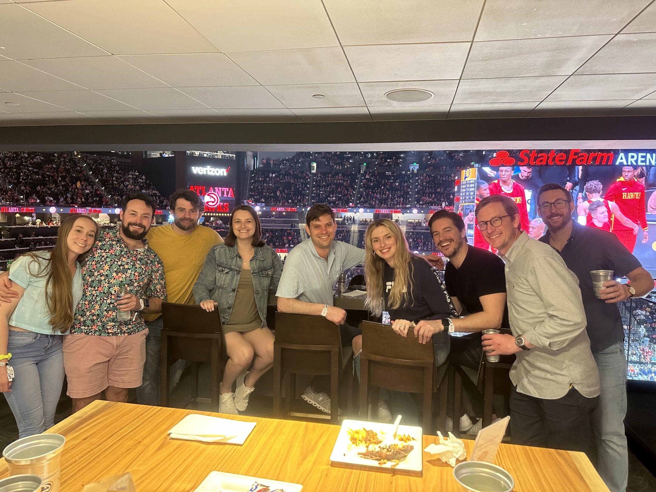 A group of nine people posing together in a suite at State Farm Arena during an Atlanta Hawks basketball game