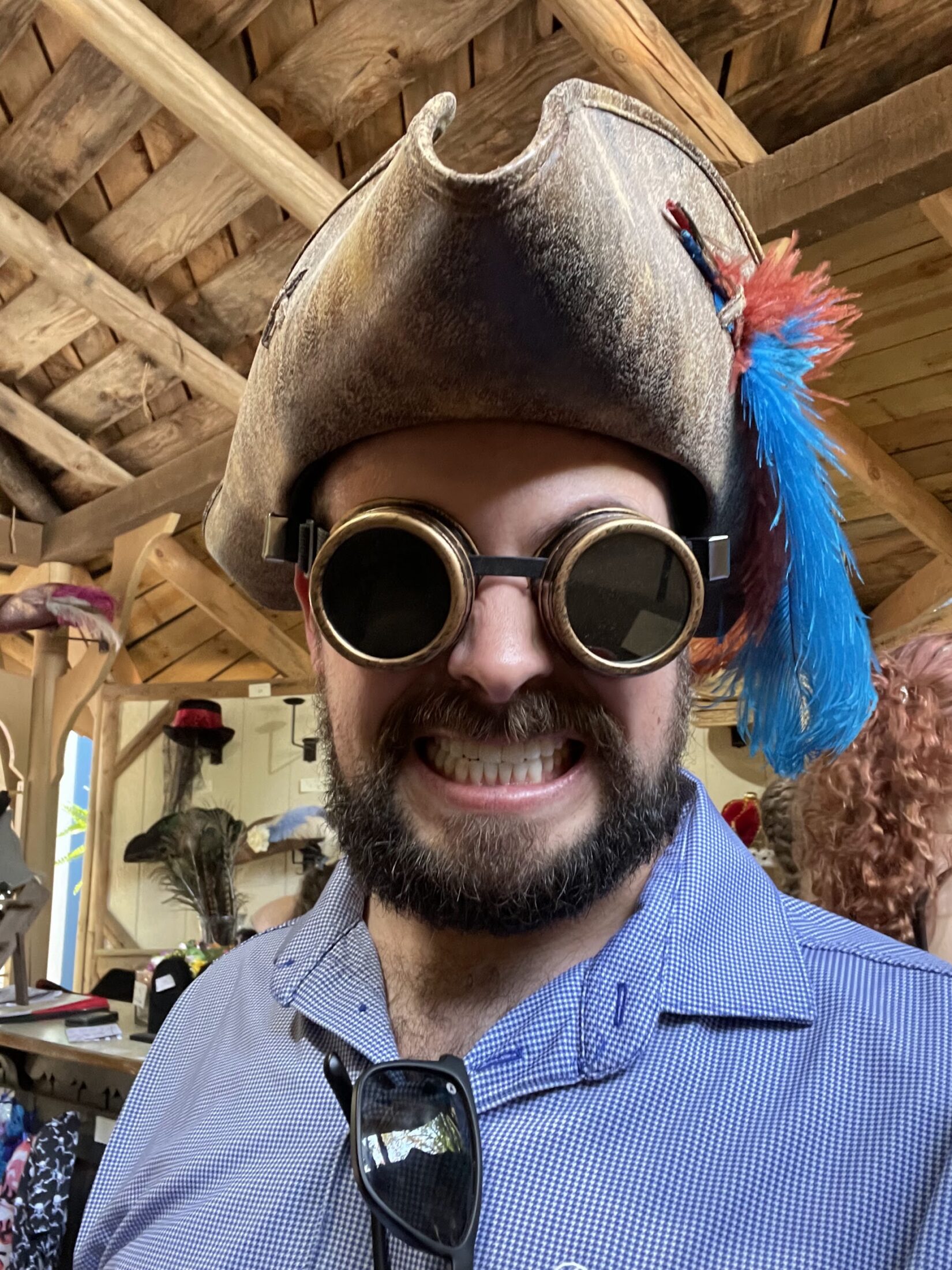 Sam Solomon wearing a pirate hat with colorful feathers and steampunk goggles, grinning inside a rustic wooden hat shop