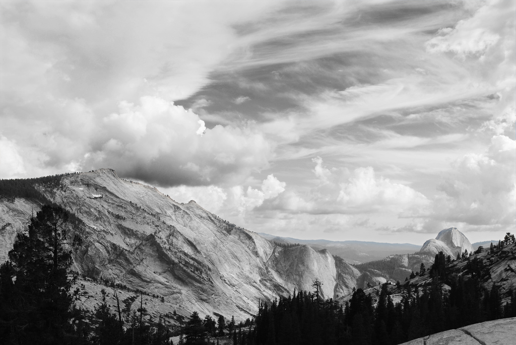 Black and white landscape of granite mountains and pine trees under dramatic swirling clouds, with Half Dome visible in the distance