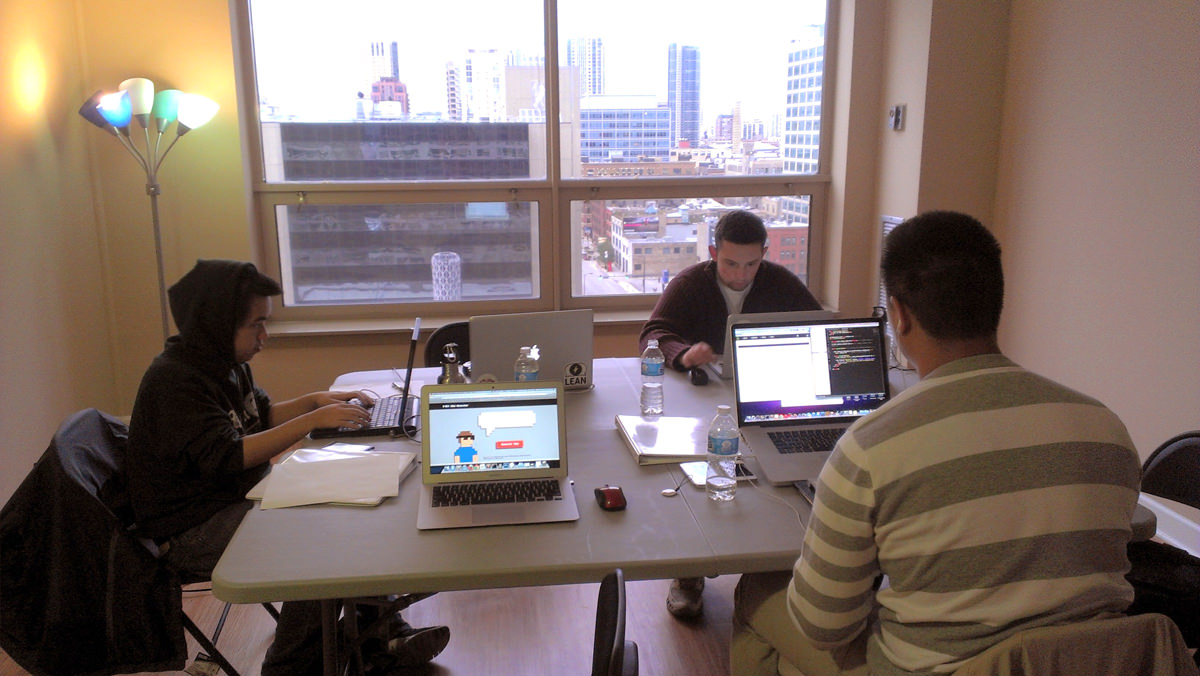 Three people working on laptops at a folding table in an office with a city skyline view through large windows